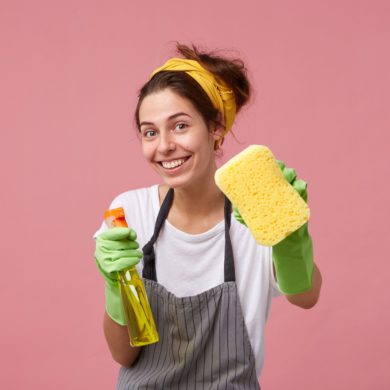 Smiling housewoman in apron and rubber gloves showing tidy sponge and detergent isolated over pink background with copy space. Beautiful joyful maid going to tidy up her room with washing spray